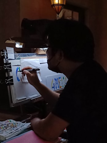 A person is seen working on a colorful piece of art in a dimly lit room. The artwork involves letters and vibrant colors, and is being created on a sheet held by clips. The environment suggests a creative workspace with a focus light and scattered art supplies.