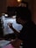 A person is seen working on a colorful piece of art in a dimly lit room. The artwork involves letters and vibrant colors, and is being created on a sheet held by clips. The environment suggests a creative workspace with a focus light and scattered art supplies.