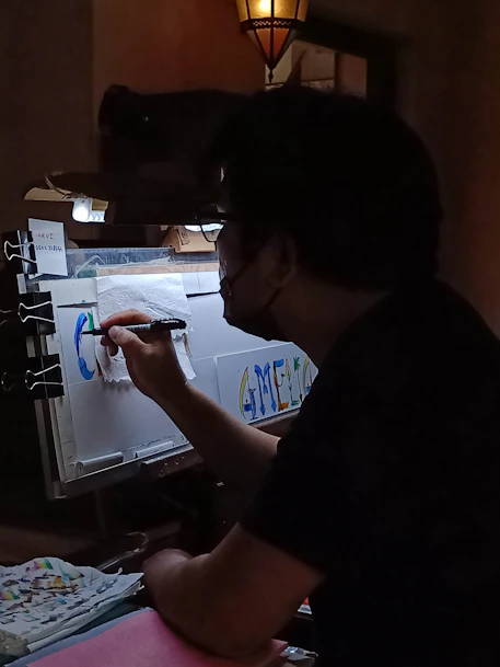 A person is seen working on a colorful piece of art in a dimly lit room. The artwork involves letters and vibrant colors, and is being created on a sheet held by clips. The environment suggests a creative workspace with a focus light and scattered art supplies.