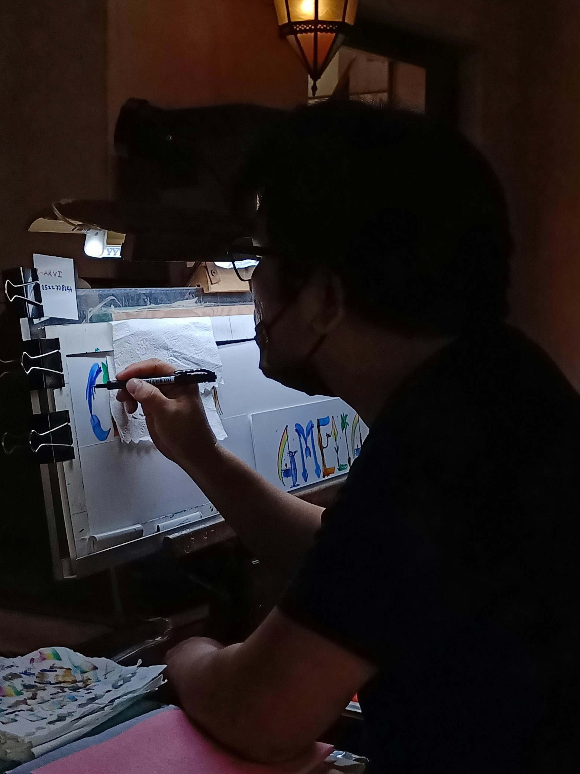 A person is seen working on a colorful piece of art in a dimly lit room. The artwork involves letters and vibrant colors, and is being created on a sheet held by clips. The environment suggests a creative workspace with a focus light and scattered art supplies.