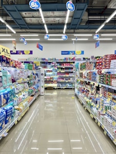 A well-organized supermarket aisle lined with various products. Shelves are neatly stocked with household items, toiletries, and packaged goods. The floor is clean and shiny, reflecting the overhead fluorescent lights. Promotional signs are hanging from the ceiling, offering discounts.