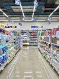 A well-organized supermarket aisle lined with various products. Shelves are neatly stocked with household items, toiletries, and packaged goods. The floor is clean and shiny, reflecting the overhead fluorescent lights. Promotional signs are hanging from the ceiling, offering discounts.