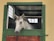 A white horse is standing inside a stable looking outside through a half-open green stable door. The background shows a corrugated metal roof and small glass windows, with sunlight gently filtering through.