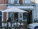 A small outdoor seating area of a restaurant with tables and chairs under a large white umbrella. There are people sitting and chatting at the tables, and a woman walking by on the sidewalk. The restaurant has a glass window with a patterned design and the name 'Pita a Pezi' is visible on the awning above.