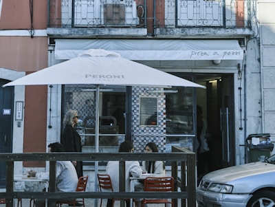 A small outdoor seating area of a restaurant with tables and chairs under a large white umbrella. There are people sitting and chatting at the tables, and a woman walking by on the sidewalk. The restaurant has a glass window with a patterned design and the name 'Pita a Pezi' is visible on the awning above.