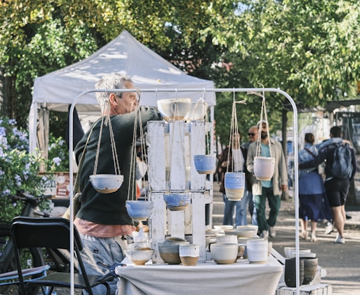 A man stands in front of a display of handmade pottery at an outdoor market. There are hanging pots and various ceramic bowls arranged on a table. People walk by in the background, and a white tent is visible, along with lush green trees and a bicycle.