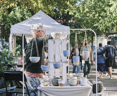A man stands in front of a display of handmade pottery at an outdoor market. There are hanging pots and various ceramic bowls arranged on a table. People walk by in the background, and a white tent is visible, along with lush green trees and a bicycle.