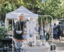 A man stands in front of a display of handmade pottery at an outdoor market. There are hanging pots and various ceramic bowls arranged on a table. People walk by in the background, and a white tent is visible, along with lush green trees and a bicycle.