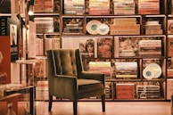 An elegant and cozy bookstore corner features a plush, green armchair set against a backdrop of neatly arranged bookshelves filled with colorful books and decorative plates. Soft lighting enhances the warm and inviting atmosphere.