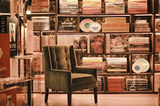 An elegant and cozy bookstore corner features a plush, green armchair set against a backdrop of neatly arranged bookshelves filled with colorful books and decorative plates. Soft lighting enhances the warm and inviting atmosphere.