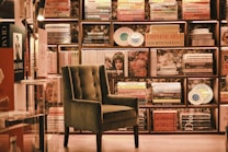 An elegant and cozy bookstore corner features a plush, green armchair set against a backdrop of neatly arranged bookshelves filled with colorful books and decorative plates. Soft lighting enhances the warm and inviting atmosphere.