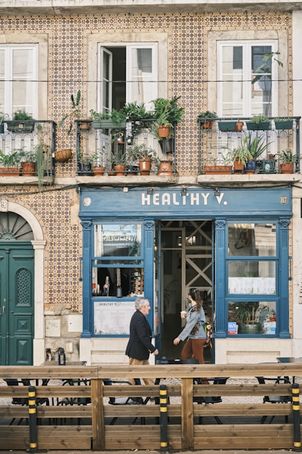 A charming building with intricate tile patterns covering the facade. The upper level features balconies adorned with various potted plants, adding a touch of greenery. The lower level has a cafe or shop with a sign reading 'HEALTHY V.' A man walks past the entrance while a woman carrying a beverage emerges from the door. Outdoor seating is visible in the foreground, separated by a wooden barrier.