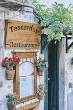 A quaint street-side restaurant with a rustic wooden sign reading 'Tascardoso Restaurante' adorned with illustrations of a pitcher and glasses. Surrounding the sign are pots of vibrant red flowers. The old stone exterior is decorated with patterned tiles, and a green door is partially visible. Overhanging branches with green leaves add to the charm.