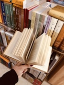 A hand holding an open book with visible text, surrounded by neatly arranged shelves filled with various books. The books have colorful spines with text, indicating a library or bookstore setting.