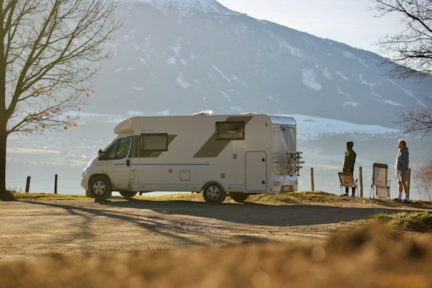 A campervan is parked on a scenic overlook, with a mountainous landscape visible in the background. Two people stand beside folding chairs, enjoying the view. Bare trees frame the scene, suggesting it is autumn or early winter.