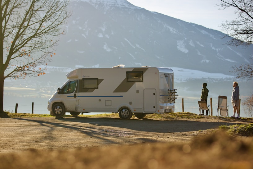 A campervan is parked on a scenic overlook, with a mountainous landscape visible in the background. Two people stand beside folding chairs, enjoying the view. Bare trees frame the scene, suggesting it is autumn or early winter.