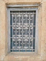 A window is enclosed by an intricate metal grille with decorative patterns. The frame and the surrounding wall appear weathered, showcasing textures of peeling paint and aged stone.