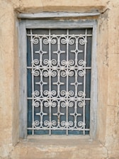 A window is enclosed by an intricate metal grille with decorative patterns. The frame and the surrounding wall appear weathered, showcasing textures of peeling paint and aged stone.
