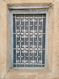 A window is enclosed by an intricate metal grille with decorative patterns. The frame and the surrounding wall appear weathered, showcasing textures of peeling paint and aged stone.