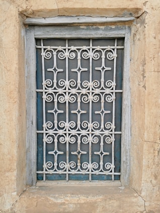 A window is enclosed by an intricate metal grille with decorative patterns. The frame and the surrounding wall appear weathered, showcasing textures of peeling paint and aged stone.