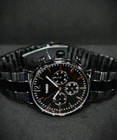 A black wristwatch with a metal link bracelet is displayed against a dark background. The watch features a round face with three smaller sub-dials, sleek metal hour and minute hands, and simple, minimalist hour markers. A brand name is visible on the watch face.