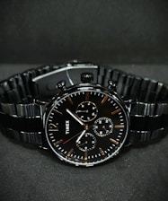 A black wristwatch with a metal link bracelet is displayed against a dark background. The watch features a round face with three smaller sub-dials, sleek metal hour and minute hands, and simple, minimalist hour markers. A brand name is visible on the watch face.
