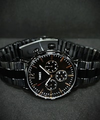 A black wristwatch with a metal link bracelet is displayed against a dark background. The watch features a round face with three smaller sub-dials, sleek metal hour and minute hands, and simple, minimalist hour markers. A brand name is visible on the watch face.
