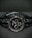 A black wristwatch with a metal link bracelet is displayed against a dark background. The watch features a round face with three smaller sub-dials, sleek metal hour and minute hands, and simple, minimalist hour markers. A brand name is visible on the watch face.