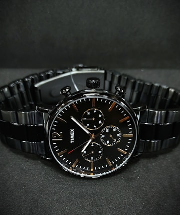 A black wristwatch with a metal link bracelet is displayed against a dark background. The watch features a round face with three smaller sub-dials, sleek metal hour and minute hands, and simple, minimalist hour markers. A brand name is visible on the watch face.