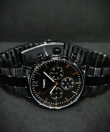 A black wristwatch with a metal link bracelet is displayed against a dark background. The watch features a round face with three smaller sub-dials, sleek metal hour and minute hands, and simple, minimalist hour markers. A brand name is visible on the watch face.