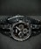 A black wristwatch with a metal link bracelet is displayed against a dark background. The watch features a round face with three smaller sub-dials, sleek metal hour and minute hands, and simple, minimalist hour markers. A brand name is visible on the watch face.