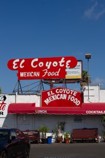 A vibrant sign displays the name 'El Coyote Mexican Food' in bold red letters with a white background. Below the sign, a red awning shades the entrance, advertising lunch and cocktails. Several potted plants and wooden benches are arranged along the white exterior of the building.