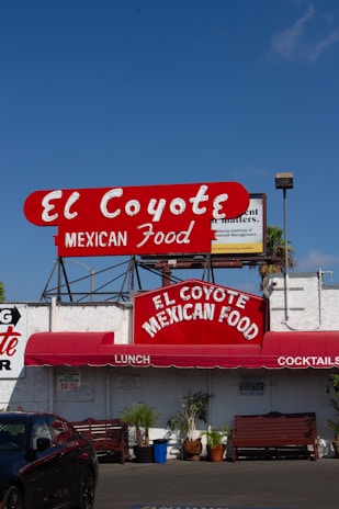 A vibrant sign displays the name 'El Coyote Mexican Food' in bold red letters with a white background. Below the sign, a red awning shades the entrance, advertising lunch and cocktails. Several potted plants and wooden benches are arranged along the white exterior of the building.