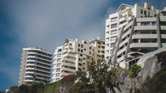 Several modern, multi-story residential buildings with a mix of geometric designs are positioned against a blue sky. The structures are surrounded by lush greenery and positioned on a slope, offering a dynamic perspective.