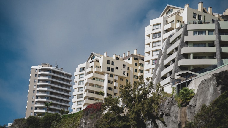 Several modern, multi-story residential buildings with a mix of geometric designs are positioned against a blue sky. The structures are surrounded by lush greenery and positioned on a slope, offering a dynamic perspective.