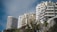 Several modern, multi-story residential buildings with a mix of geometric designs are positioned against a blue sky. The structures are surrounded by lush greenery and positioned on a slope, offering a dynamic perspective.