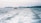 A small boat travels across the ocean, creating a foamy wake behind it. The sea is vast and expansive, with distant hills or islands visible on the horizon under a cloudy sky.