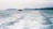 A small boat travels across the ocean, creating a foamy wake behind it. The sea is vast and expansive, with distant hills or islands visible on the horizon under a cloudy sky.