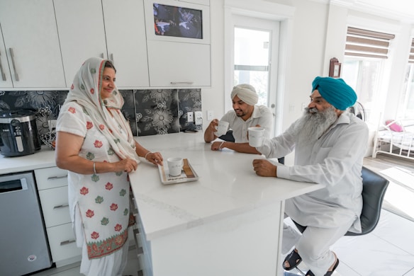 Three people are gathered in a modern kitchen, engaging in conversation and holding mugs. The woman is wearing a traditional dress with floral patterns and a headscarf, while two men are wearing turbans. The kitchen has white cabinetry and modern appliances.