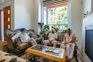 Three individuals are seated in a living room, engaged in conversation. They are dressed in traditional attire and sitting on ornate sofas. Large windows allow natural light to illuminate the room, which is decorated with plants and a fireplace.