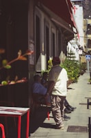 A group of people sit and stand along a sidewalk, shaded by a building's awning. Small tables and chairs are positioned along the path, and red accents are highlighted against the neutral concrete and building facade. There are potted plants lining the walkway, and the scene suggests a peaceful social setting in an urban environment.