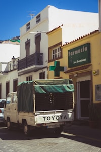A narrow street features a white Toyota truck with a green tarp covering its rear parked in front of a building. The building on the right has a sign for a pharmacy named 'Farmacia San Bartolomé'. The architecture is traditional with barred windows and a wrought iron balcony. The scene is well-lit, indicating a sunny day.