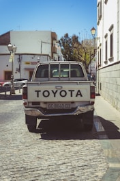 A white Toyota pickup truck is parked on a cobblestone street in an urban area. Surrounding the vehicle are historic-style street lamps and buildings, with one building displaying a sign. Behind the truck, there is a mix of modern and older architecture, including a row of leafless trees.