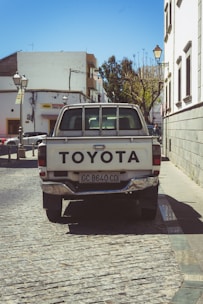 A white Toyota pickup truck is parked on a cobblestone street in an urban area. Surrounding the vehicle are historic-style street lamps and buildings, with one building displaying a sign. Behind the truck, there is a mix of modern and older architecture, including a row of leafless trees.