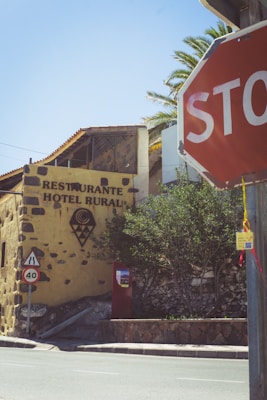 A rustic stone building with signage for a rural hotel and restaurant. The facade features a mix of stone and stucco with visible text and geometric designs. There is a stop sign prominently positioned in the foreground and a speed limit sign indicating 40 km/h nearby. The surrounding area includes lush green trees set against a clear blue sky.