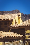A rustic building with a yellow facade and clay tile roofing, set against a backdrop of rocky mountains under a clear blue sky. The architecture reflects a traditional style with a chimney visible atop one of the roofs.