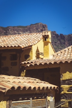 A rustic building with a yellow facade and clay tile roofing, set against a backdrop of rocky mountains under a clear blue sky. The architecture reflects a traditional style with a chimney visible atop one of the roofs.