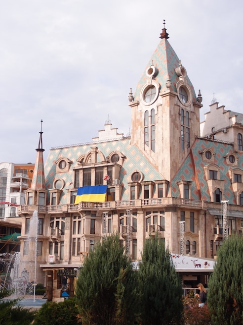 A grand building with intricate architectural features and tall, pointed roofs, adorned with large round windows. The structure displays a colorful tiled pattern on its roof and features a Ukrainian flag hanging prominently. In the foreground, several lush green bushes and a fountain can be seen, with water spraying upwards.