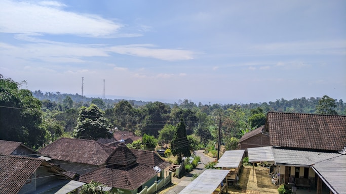 A rural landscape featuring a cluster of houses with tiled roofs surrounded by lush green trees. In the background, there are rolling hills and two visible communication towers. A narrow dirt road winds through the community, indicating a peaceful village setting under a partly cloudy sky.