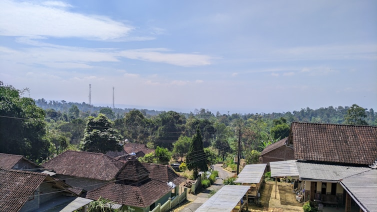 A rural landscape featuring a cluster of houses with tiled roofs surrounded by lush green trees. In the background, there are rolling hills and two visible communication towers. A narrow dirt road winds through the community, indicating a peaceful village setting under a partly cloudy sky.